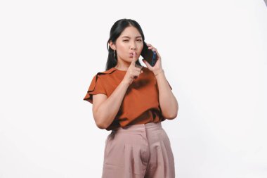 Güzel, Asyalı bir kadın, beyaz arka planla izole edilmiş akıllı telefonu tutarken sessiz ya da gizlice işaret eden kahverengi tişört giyiyor..