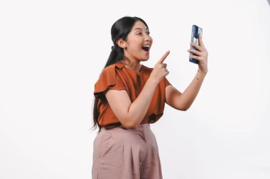 Kahverengi tişörtlü neşeli genç Asyalı kadın elinde tuttuğu cep telefonunu işaret ediyor mutlu bir gülümsemeyle, izole edilmiş beyaz arka planla..