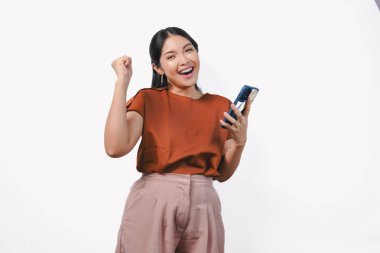 Heyecanlı genç Asyalı kadın, beyaz arka planla izole edilmiş akıllı telefonu tutarken mutlu bir ifadeyle yumruklarını kaldırıyor..