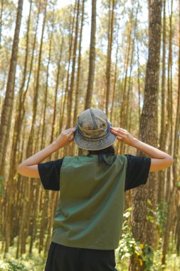 Back view of a young Asian woman wearing a green vest and hat standing in a forest and raising her hands in the air.