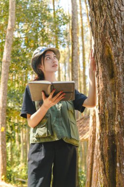 Beautiful young Asian woman is holding a book while studying the tree and nature in the forest.