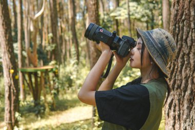Pretty young Asian woman wearing green vest and hat is holding a camera, taking picture of the forest nature.