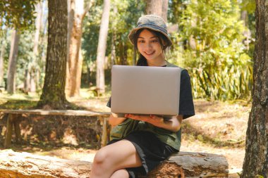 Yeşil yelek ve şapkalı gülümseyen Asyalı kadın ormanda kamp yaparken laptopunu kullanıyor..