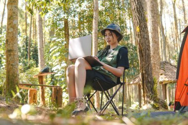Yeşil yelek ve şapkalı gülümseyen Asyalı kadın ormanda kamp yaparken laptopunu kullanıyor..