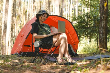 Yeşil yelek ve şapka giyen neşeli genç Asyalı kadın çadırın dışında bir sandalyede otururken laptopunu kullanıyor..