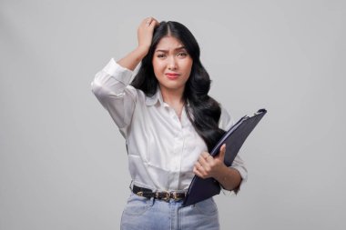 Beyaz gömlekli ve kot pantolonlu kafası karışmış genç bir Asyalı kadın elinde bir belge panosu tutarken stresli, beyaz arka planda izole edilmiş..