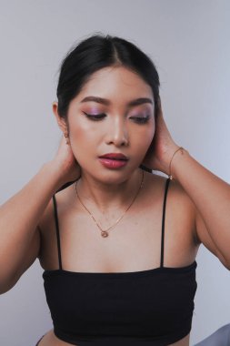 Zarif genç Asyalı kadın kameraya poz veriyor. Makyajlı. Saçını arkaya doğru tutuyor..