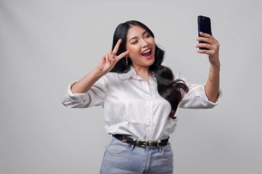 Gülümseyen genç Asyalı kadın cep telefonuyla selfie çekiyor. Barış parmağı hareketleriyle izole edilmiş beyaz arka plan üzerinde..