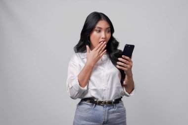 Beyaz tişörtlü genç Asyalı kadın elinde akıllı bir telefon beyaz arka planla izole edilmiş, yüzünde şaşırmış bir ifadeyle..