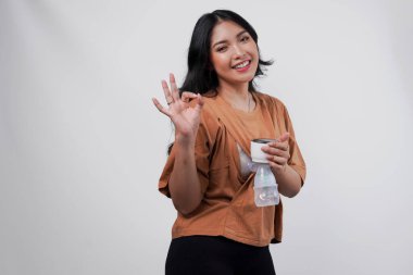 Gülümseyen Asyalı anne beyaz arka planla izole edilmiş parmak hareketi yaparken otomatik göğüs pompası ile anne sütü pompalıyor..