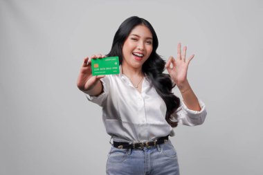 Heyecanlı genç Asyalı kadın kredi kartı ya da banka kartı sunarken beyaz arka planla izole edilmiş parmak hareketi yapıyor..