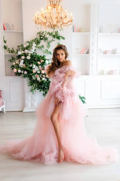 stock image Young brunette girl with long beautiful hair in a lush pink tulle dress in a light white studio on the background of shelves with books, a fireplace and flower arrangements. High quality photo