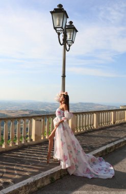 Şık bir elbiseyle dalgalı saçlı genç bir esmer, büyük bir tren ve gün batımında bir gözlem güvertesinde çiçek desenli, dağları ve denizi gören. Kaliteli fotoğraf