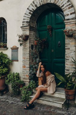 Genç bir kız avlusu yeşil bir kapının eşiğinde uzun düz saçlı çalılarla, şortlu ve gömlekli bej bir çıplak takım elbiseyle oturuyor. Yüksek kalite fotoğraf