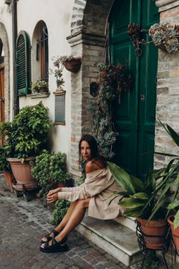 Genç bir kız avlusu yeşil bir kapının eşiğinde uzun düz saçlı çalılarla, şortlu ve gömlekli bej bir çıplak takım elbiseyle oturuyor. Yüksek kalite fotoğraf