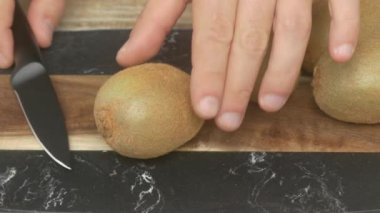 Keskin bir bıçakla olgun bir kivi meyvesini kırsal bir tahtada dikkatlice dilimleyen ellerin yakın plan görüntüsü.