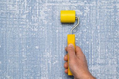 Canlı sarı bir boya silindirini dokulu mavi bir duvara sıkıca saran bir el, yenileme temaları için mükemmel.