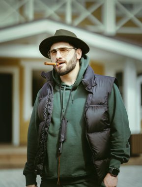 Sakallı, puro içen yakışıklı bir adamın portresi. Kovboy şapkalı genç beyaz adam evin önünde puro içiyor..