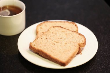 Arka planda bir fincan çay olan küçük beyaz bir tabakta iki dilim tam tahıllı ekmek. Makro fotoğrafçılık. Siyah granit arka plan.