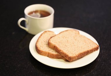 Arka planda bir fincan çay olan küçük beyaz bir tabakta iki dilim tam tahıllı ekmek. Makro fotoğrafçılık. Siyah granit arka plan.
