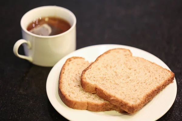 Two slices of whole grain bread on a small white plate with a cup of tea in the background. Macro photography. Black granite background.
