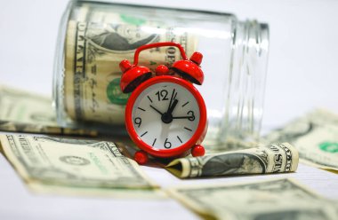 Ahşap bir masanın üzerinde cam kavanozlu dolar banknotları ve kompozisyonda kırmızı bir çalar saat. Para tasarruf kavramı.