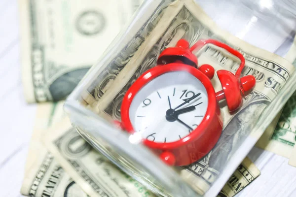 stock image Dollar bills with glass jar on a wooden table with a red alarm clock in the composition. Money saving concept.