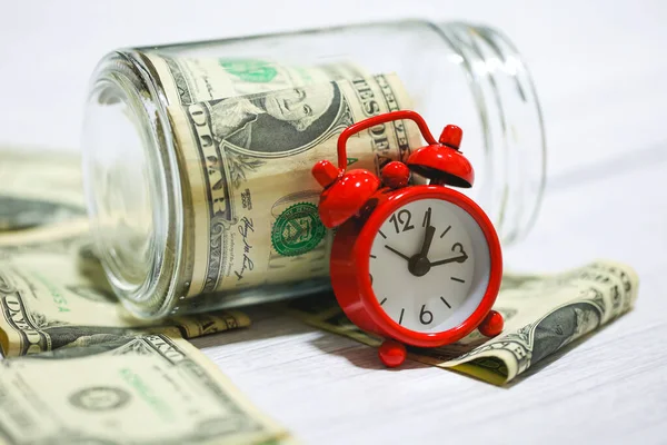 Stock image Dollar bills with glass jar on a wooden table with a red alarm clock in the composition. Money saving concept.