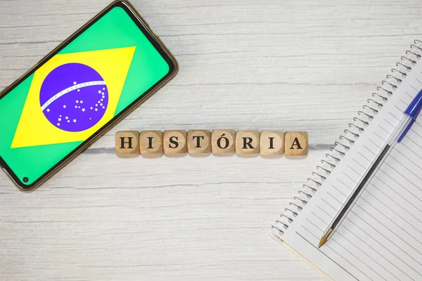 木製のテーブルの上に木製の立方体に書かれたポルトガル語のテキスト履歴 組成物中のブラジルの旗を持つノートブック 携帯電話 — ストック写真
