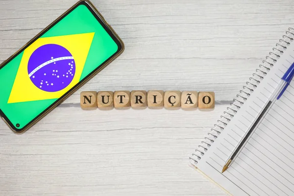 stock image The text NUTRITION in Portuguese written on wooden cubes on a wooden table. A notebook, a pen and a cell phone with the Brazilian flag in the composition. 