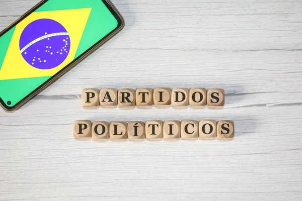 The text POLITICAL PARTIES in Brazilian Portuguese written on wooden cubes. A cell phone with the Brazilian flag being shown on the screen in the composition.