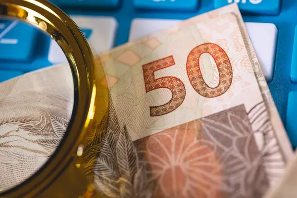 stock image Brazilian Real banknotes and a golden magnifying glass.Brazilian economy, finance and income tax.