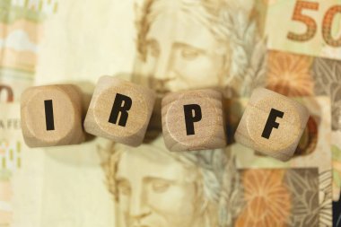 The abbreviation IRPF for personal income tax written on wooden dice. Brazilian Portuguese. Brazilian Real 50 Reais notes. Income tax. clipart
