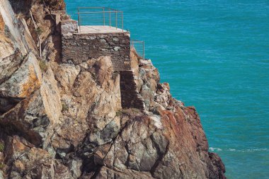 Sahildeki bir uçurumda merdivenler. Gözlem güvertesi. Mavi denizde taşlar, kayalık plajlar, yazın günbatımında dalgalar Liguria, İtalya 'da. Tropikal arka plan. Şeffaf gök mavisi su ve taşlar.