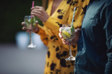 Kadın ve erkeğin elleri kadehlerle tezahürat yapar. Düğünü kutlayan bir çift, kokteyllerle yıldönümü, limonlu ve yeşillikli alkollü içecek. Yaz içkileri, düğün, parti.