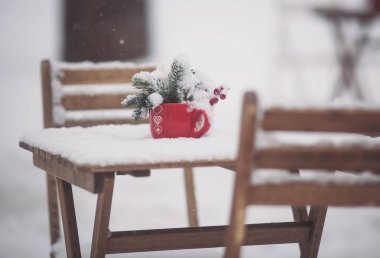 Kırmızı fincanda ladin dalları ve karlı ahşap masa üzerinde kırmızı böğürtlen var. Masası, sandalyeleri ve Noel süslemeleri olan bir Noel terası. Karlı bir kış sokak sahnesi. Noel havası.
