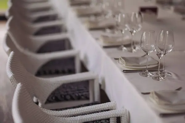 Table setting for a party or wedding reception in a restaurant. Row of ladder-back chairs set around elegant dining table with white tablecloth and crystal glasses.