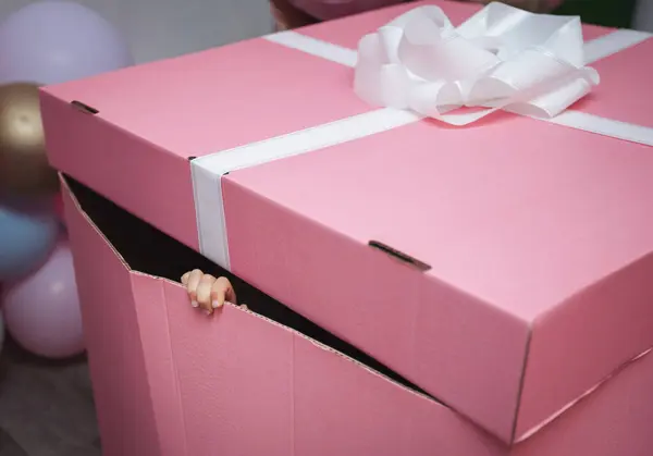 Large pink gift box with white ribbon and bow, kid\'s fingers peeking out of the box. Happy Baby inside of a gift box.
