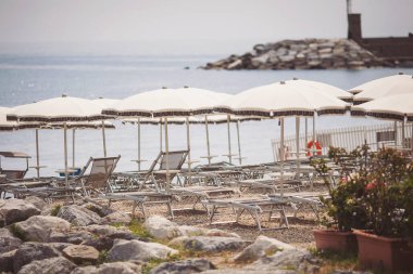 Boş İtalyan plajı. Akdeniz kıyıları, boş güneş yatakları, güneşten şemsiyeler, dağlar, çakıl taşları. Otel şezlonglarında plaj şemsiyeleri. seyahat.