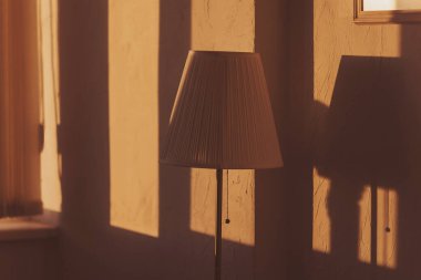 Sabah lambası ve oda duvarında güneş ışığı gölgesi. Sabah vakti sıcak bir atmosfer. Bir odanın içinde lamba, ışık ve gölge. Odada klasik lamba direği var. Duvarda asgari biçim ve gölge deseni