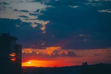 Şehirde kırmızı ve mavi gün batımı. Ateşli şafağı olan şehir manzarası. Şehir binalarının karanlık siluetlerinin üzerindeki inanılmaz sıcak, dramatik bulutlu gökyüzü. Turuncu güneş ışığı. Bulutlu havada gün doğumu.