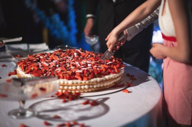 Gelin ve damat büyük bir düğün pastası kesiyor. Üstünde çilekler var..