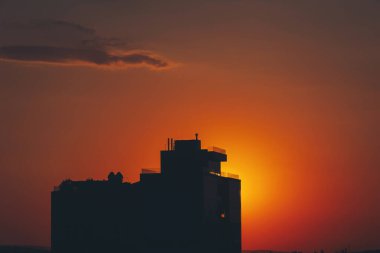 Göz kamaştırıcı bir günbatımı, güneş bulutların içine inerken ön planda uzak bir binayla gökyüzünü boyar. Sahne şehir ve doğal elementlerin güzelliğini yakalıyor, alacakaranlıkta uyum içinde harmanlıyor..