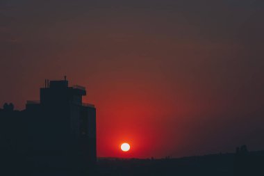 Göz kamaştırıcı bir günbatımı, güneş bulutların içine inerken ön planda uzak bir binayla gökyüzünü boyar. Sahne şehir ve doğal elementlerin güzelliğini yakalıyor, alacakaranlıkta uyum içinde harmanlıyor..