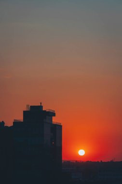 Göz kamaştırıcı bir günbatımı, güneş bulutların içine inerken ön planda uzak bir binayla gökyüzünü boyar. Sahne şehir ve doğal elementlerin güzelliğini yakalıyor, alacakaranlıkta uyum içinde harmanlıyor..