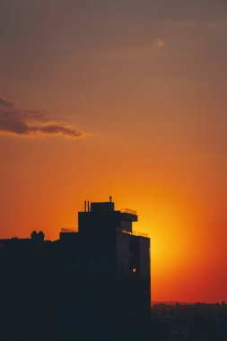 Göz kamaştırıcı bir günbatımı, güneş bulutların içine inerken ön planda uzak bir binayla gökyüzünü boyar. Sahne şehir ve doğal elementlerin güzelliğini yakalıyor, alacakaranlıkta uyum içinde harmanlıyor..