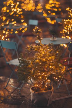 Christmas tree in pot on city street near cafe. Restaurant terrace decorating potted xmas trees.  Focus on the Christmas tree in the foreground. lights bokeh. clipart