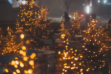 Christmas tree in pot on city street near cafe. Restaurant terrace decorating potted xmas trees.  Focus on the Christmas tree in the foreground. lights bokeh. clipart