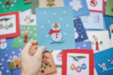 Hand holding quilling card with snowman with red scarf looks up at falling snow. woman making greeting cards. Hand made of paper quilling technique. Handicraft at home. Hobby, home office. clipart