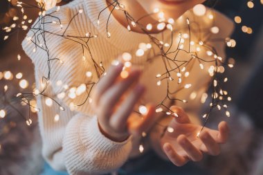 Yakın plan, birbirine dolanmış çelenkler küçük kız ellerinde, üst manzara. Elinde Noel ışıkları olan beyaz kazaklı bir kız, sıcak Noel havasında.